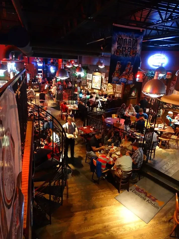 Lively, multi-level restaurant interior with patrons, Mardi Gras banner, and industrial-rustic decor.