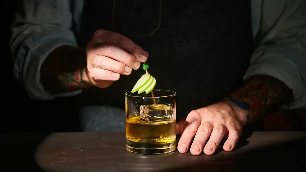 Close-up: tattooed hands garnishing an amber cocktail with apple slices.