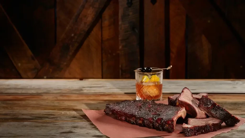 Dark, smoky ribs and an amber cocktail on a rustic wooden table.