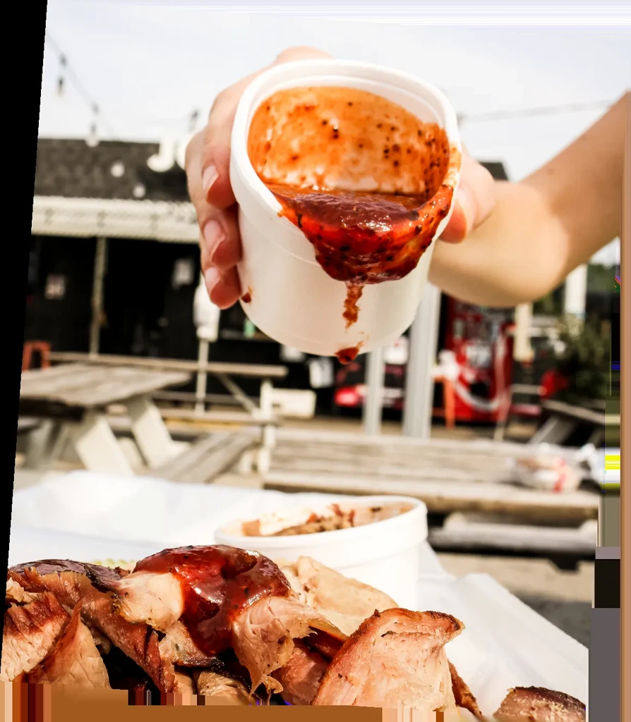 Hand pouring chunky BBQ sauce onto pulled pork outdoors.