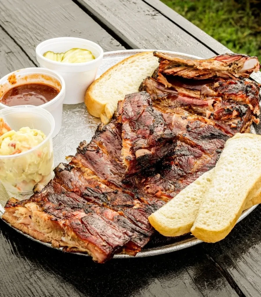 Platter of barbecued ribs with bread, pickles, sauce, and creamy salad.