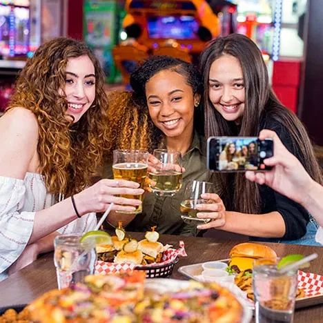 Smiling women toast drinks and food, an arcade behind, selfie visible.