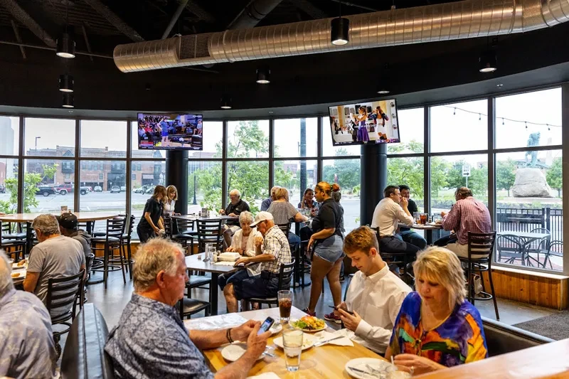 Lively modern restaurant interior with industrial decor, city view, and many diners.