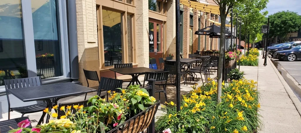 Sunny outdoor patio with black tables, chairs, flowers, and umbrellas.