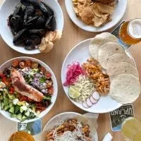 Overhead flat lay of diverse dishes: mussels, tacos, salmon salad, beer.