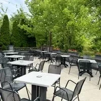 Empty patio with light tables, dark chairs, green trees, string lights.