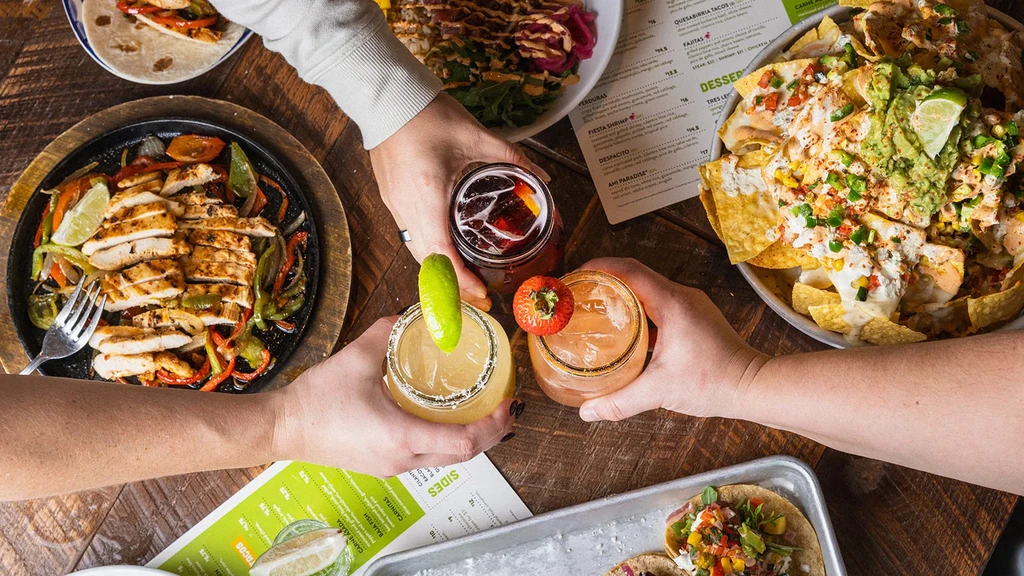Hands toasting drinks above Mexican fajitas, nachos, and tacos.