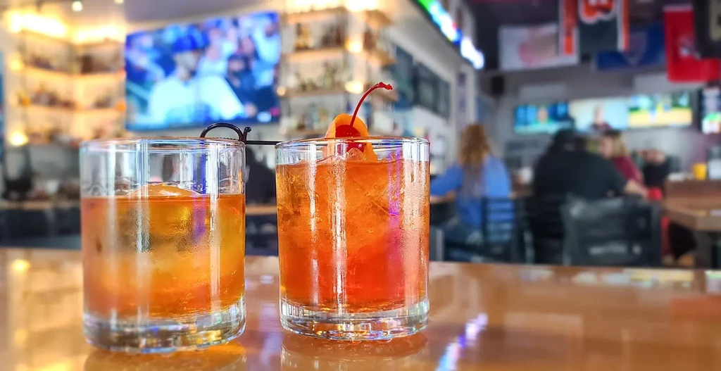 Two garnished amber cocktails on a bar counter with a blurred sports bar background.