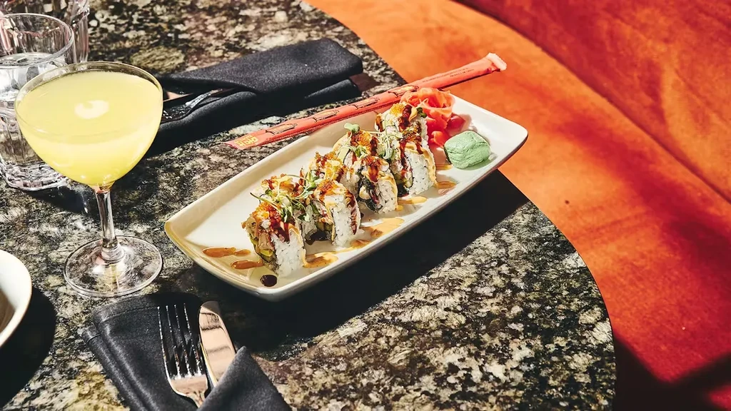 Garnished sushi rolls, yellow cocktail, and chopsticks on a dark stone table.