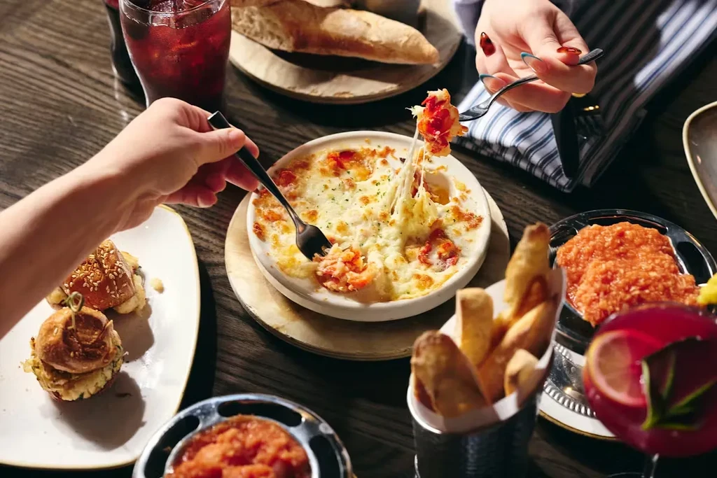Two hands pull stringy melted cheese from a baked dish with seafood.