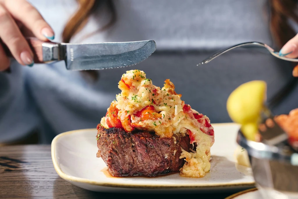 Close-up: Hands poised to cut steak topped with lobster meat.