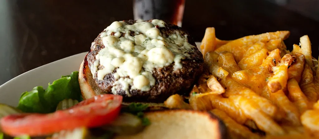 Close-up of a grilled blue cheese burger with cheesy fries and gherkins.