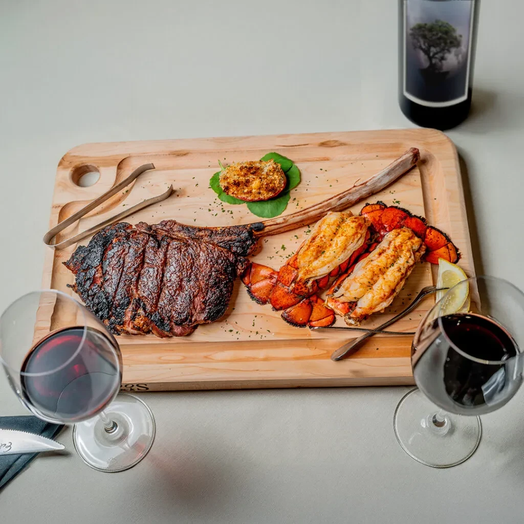 Seared tomahawk steak, grilled lobster tails, red wine on a wooden board.