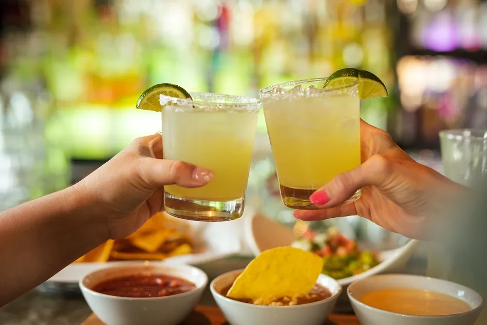Two hands clinking margarita glasses, with dips and chips.
