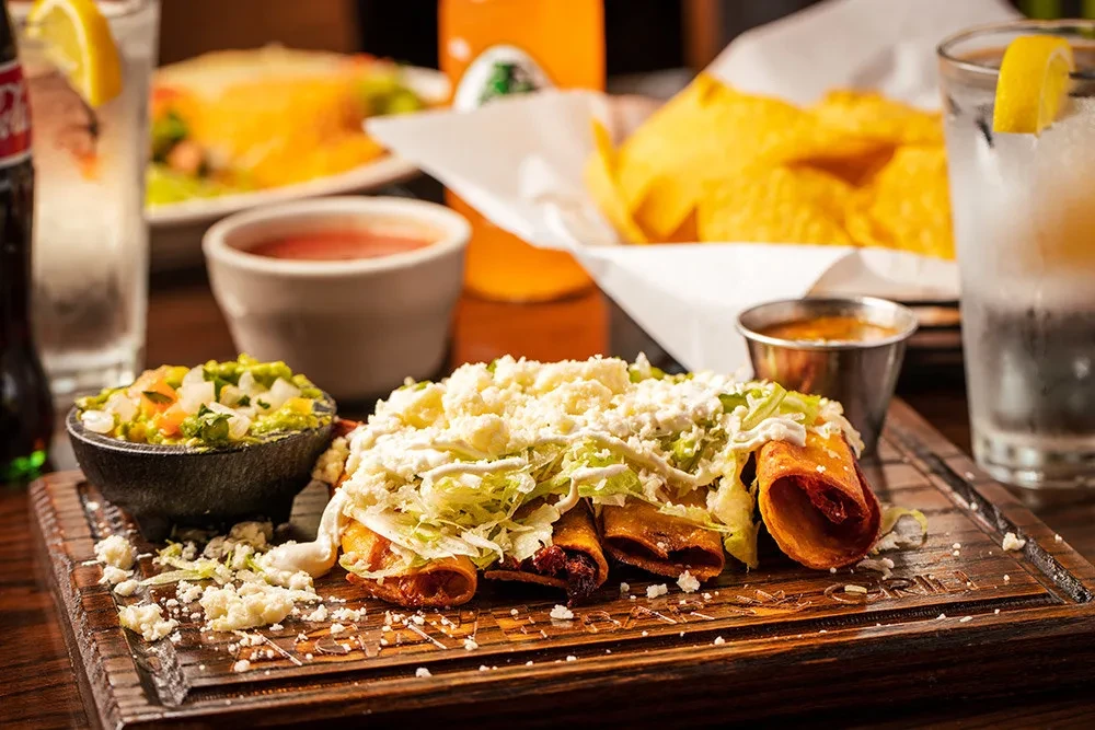 Golden taquitos with lettuce, crema, cheese, and guacamole on a board.