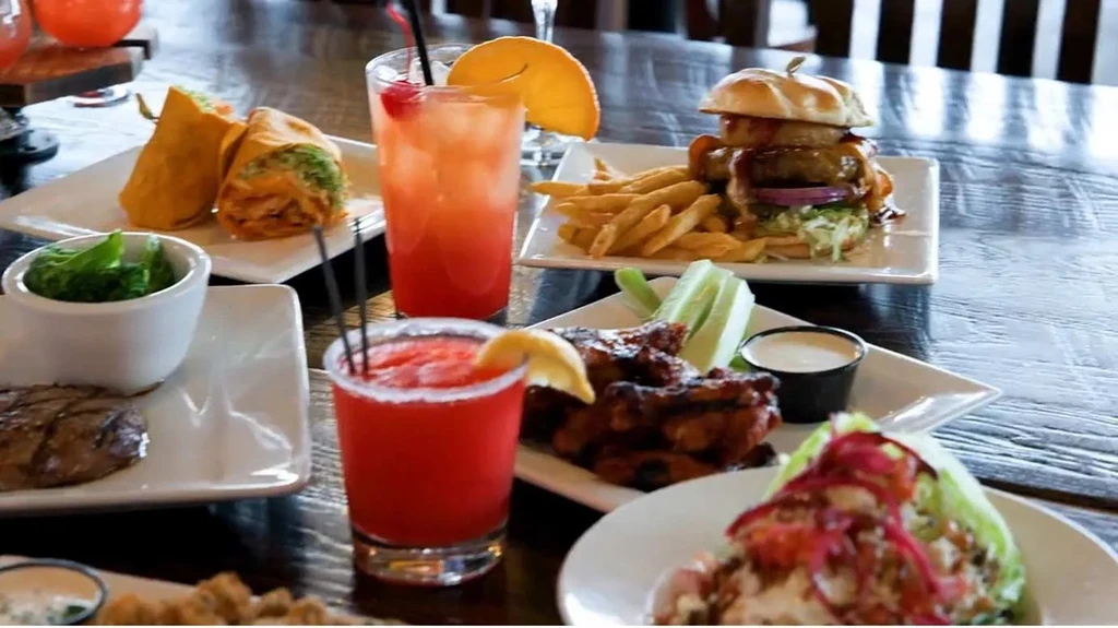 Burger, wings, steak, burritos, and cocktails on a dark wooden table.