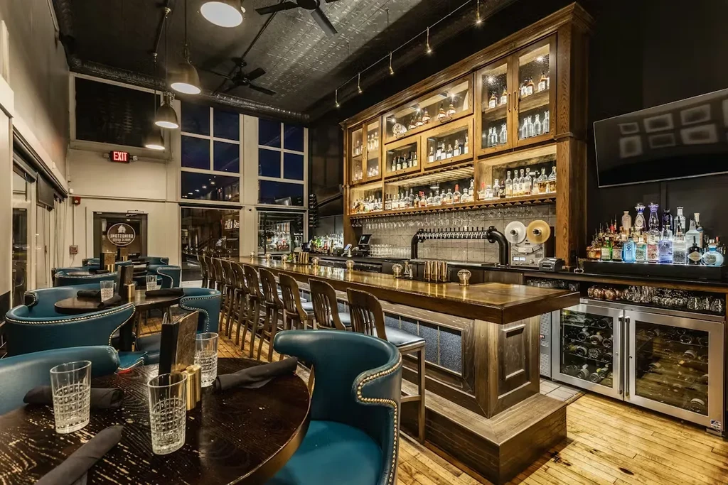 Modern restaurant interior: tables, teal chairs, and a well-stocked bar.