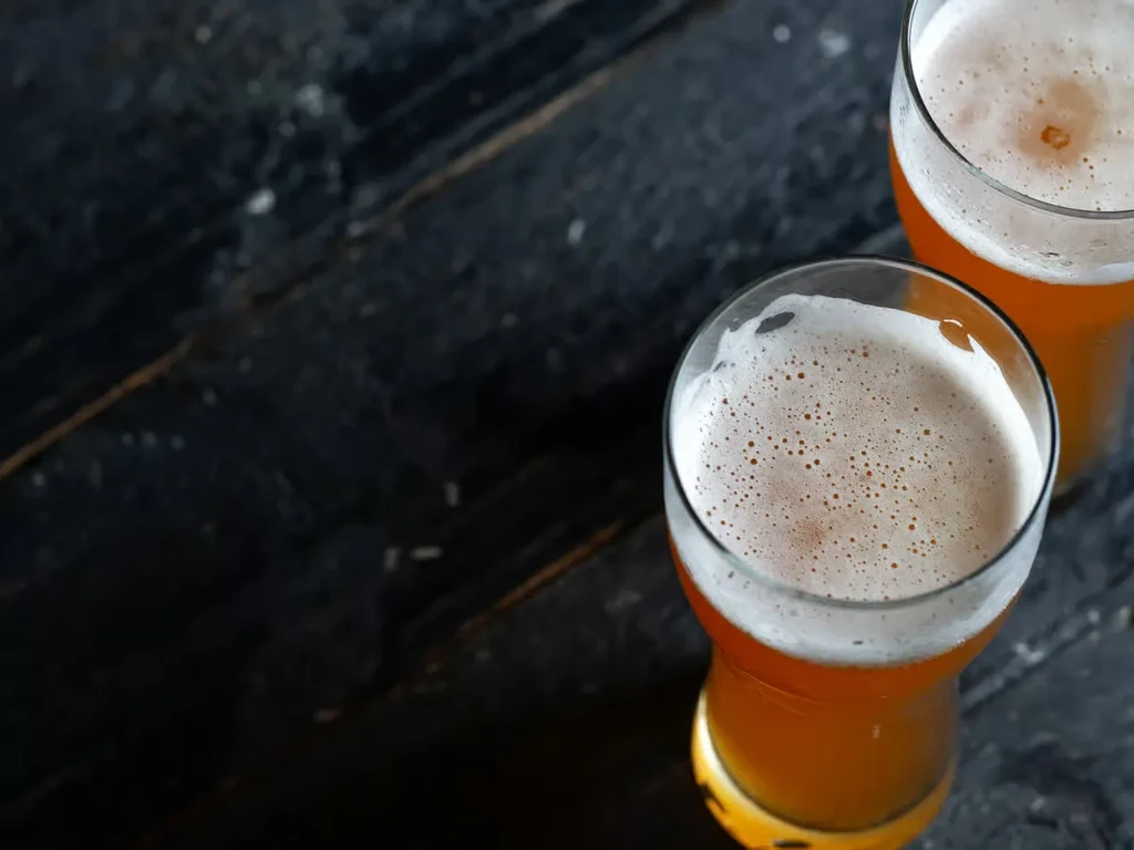 Top-down: two golden amber beers with frothy foam on dark surface.