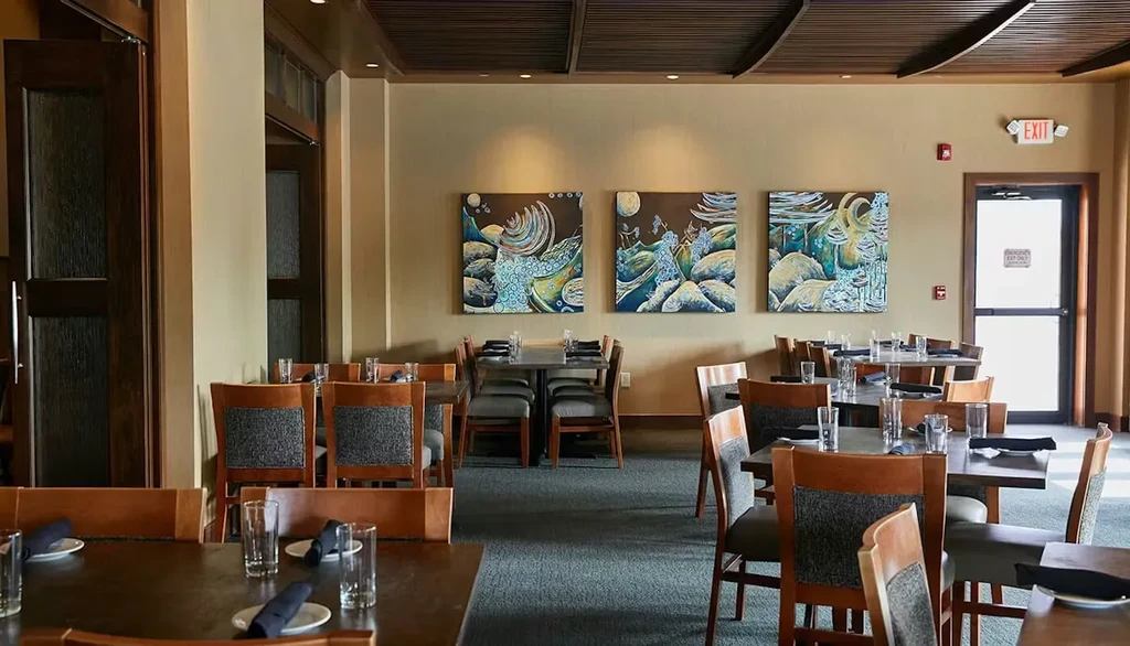 Empty restaurant dining room with dark tables, grey chairs, abstract paintings.