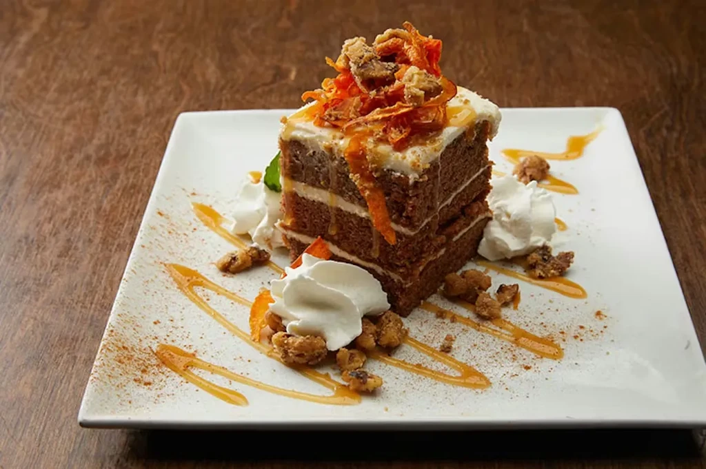 Elegant carrot cake slice with candied toppings, nuts, whipped cream, and sauce.