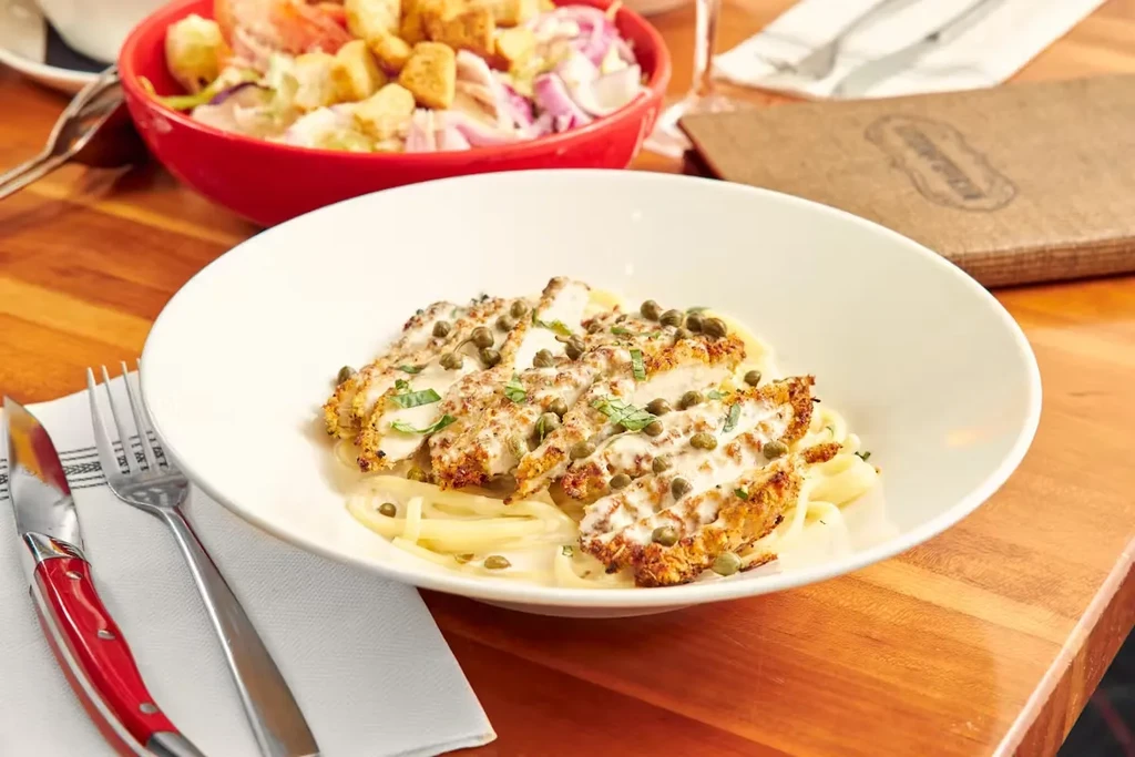 Close-up: pasta with breaded chicken, creamy sauce, capers, herbs, and salad.