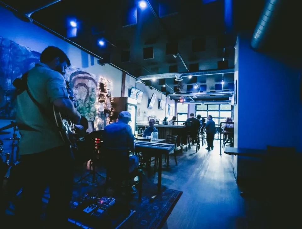 Musicians play acoustic guitar and keyboard in a blue-lit bar.