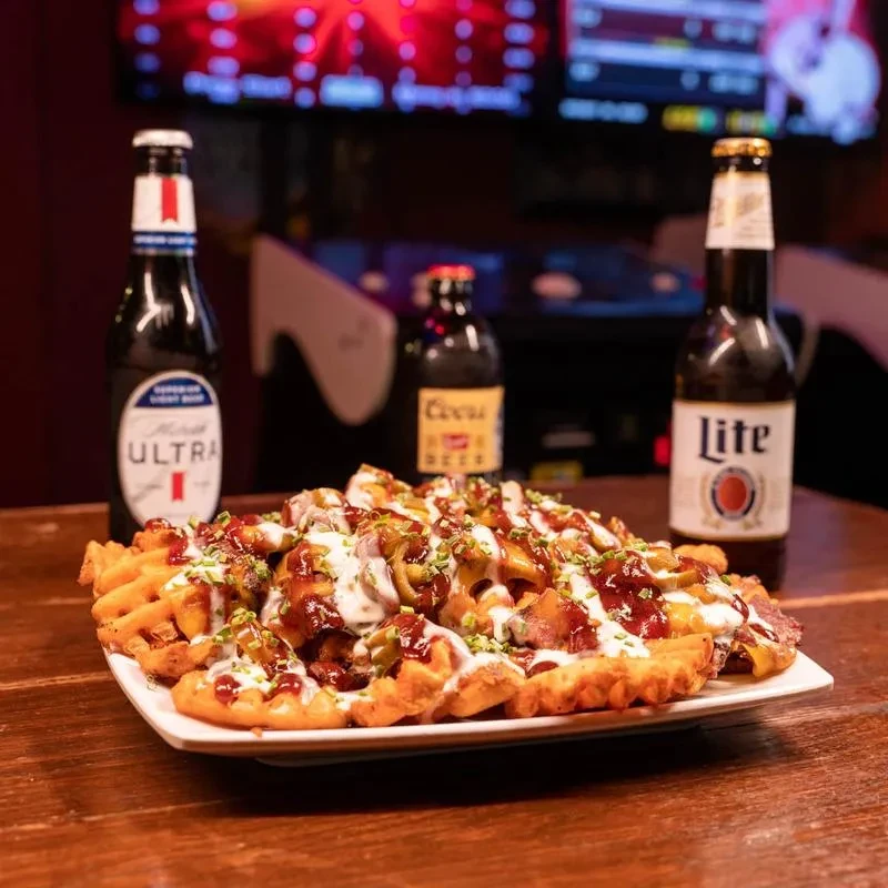 A plate of loaded waffle fries with beer bottles at a bar.