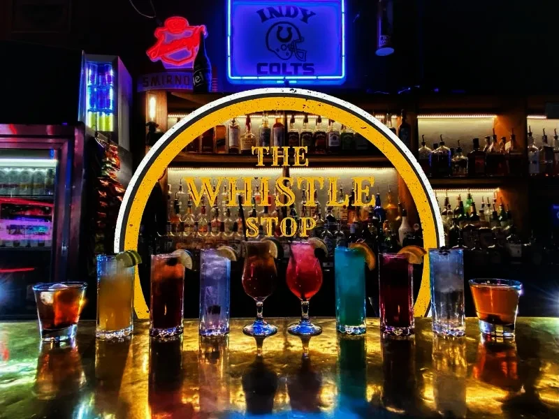 Nine colorful cocktails on a reflective bar with "THE WHISTLE STOP" sign.
