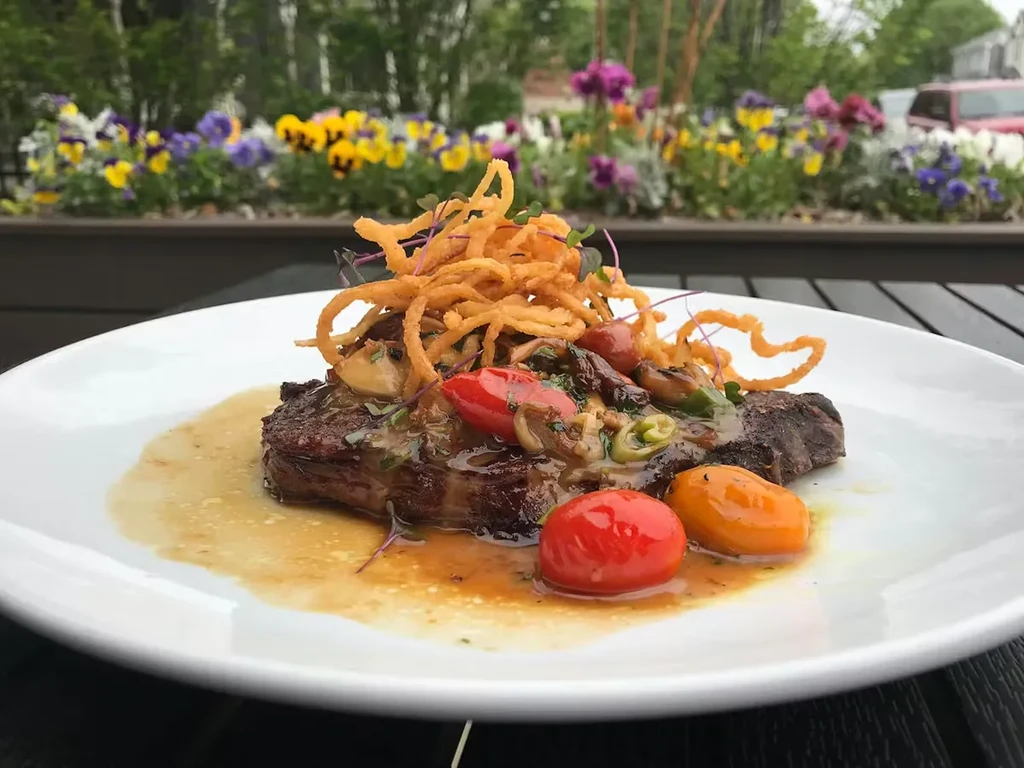 Close-up of steak with crispy onions, mushrooms, tomatoes outdoors.