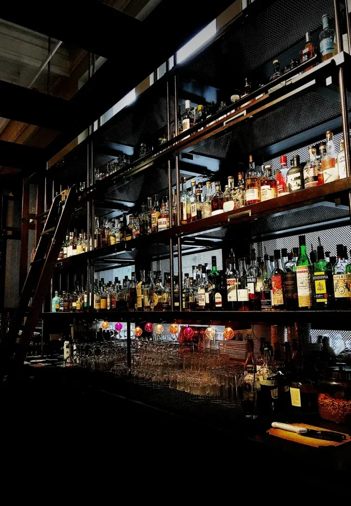 Dimly lit bar with tall liquor shelves, a ladder, and colorful lights.