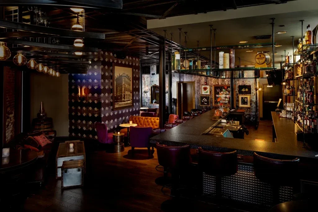 Dark bar interior with glowing liquor bottles and patterned lounge area.