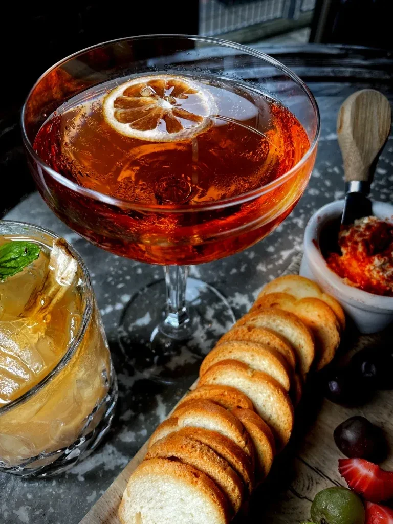 Close-up of cocktails, baguette, dip, and fruit on a wooden board.