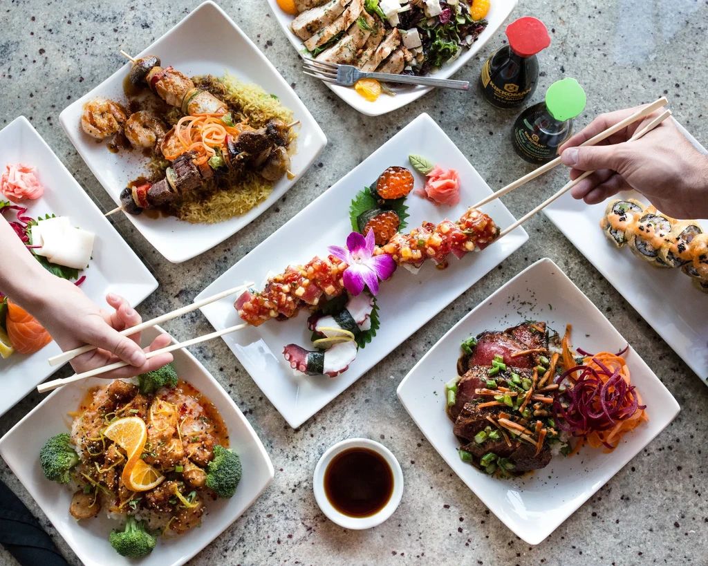 Restaurant table with diverse food: sushi, skewers, and hands using chopsticks.