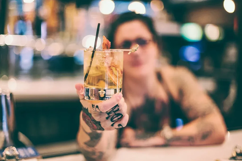 Close-up of a tattooed hand holding a garnished amber cocktail.