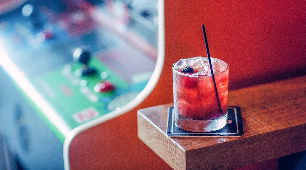Deep pink cocktail with berries and ice; blurred vintage arcade background.