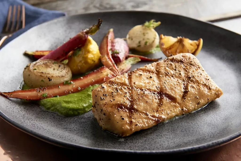 Grilled fish/chicken, colorful roasted vegetables, green puree, lemon on dark plate.