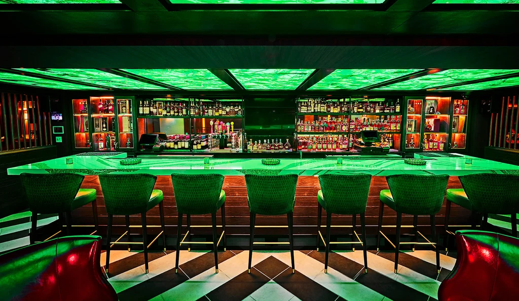 Green-lit bar with liquor bottles, geometric floor, and red chairs.