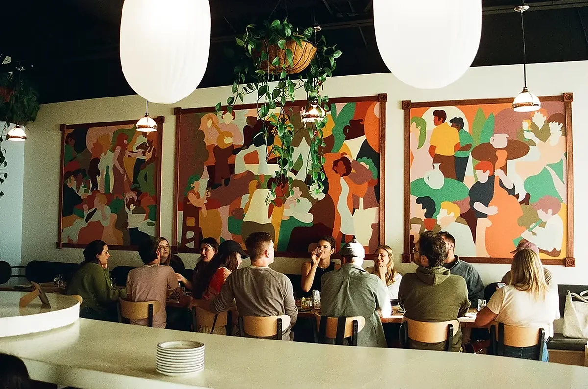 Guests dining at long table beneath colorful murals and globe pendant lights