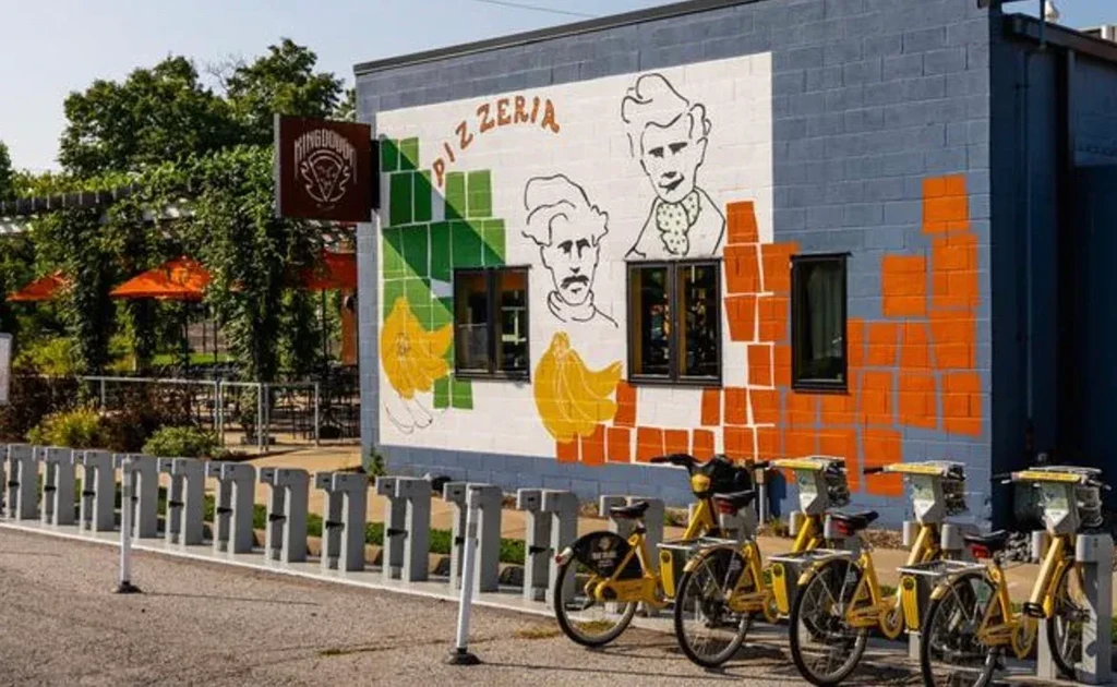 Vibrant pizzeria facade with 'PIZZERIA' mural, patio umbrellas, yellow bikes.