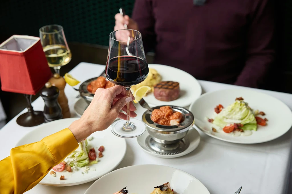 Hand holding red wine at a table with steak, salad, and seafood.