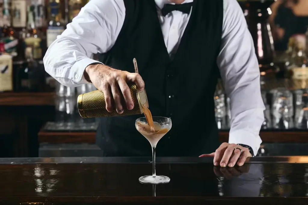 Bartender pours dark amber cocktail from golden shaker into stemmed glass.