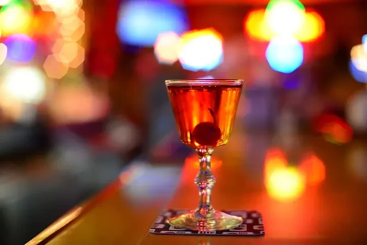 Elegant cocktail glass with cherry on bar counter, vibrant blurred lights.