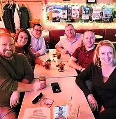 Six smiling adults with drinks in a reddish, warmly lit bar.