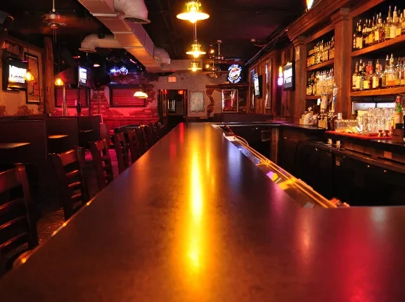 Warmly lit bar interior, long reflective counter, stocked shelves, brick walls.