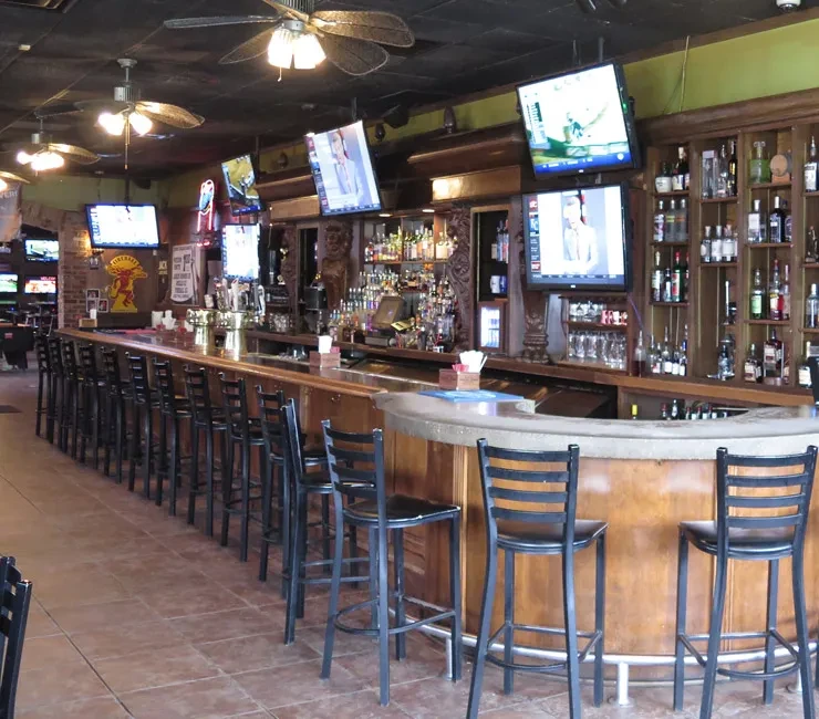 Unoccupied bar interior with curved counter, liquor bottles, and TVs.