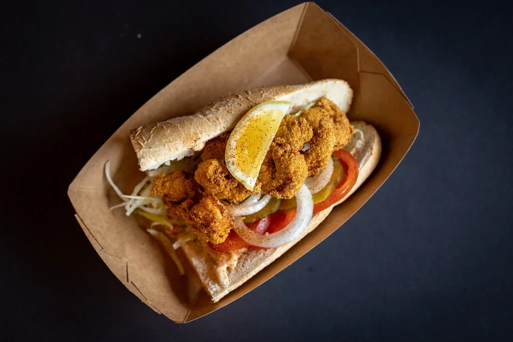 Close-up of a fried seafood po'boy with lemon in a takeout tray.