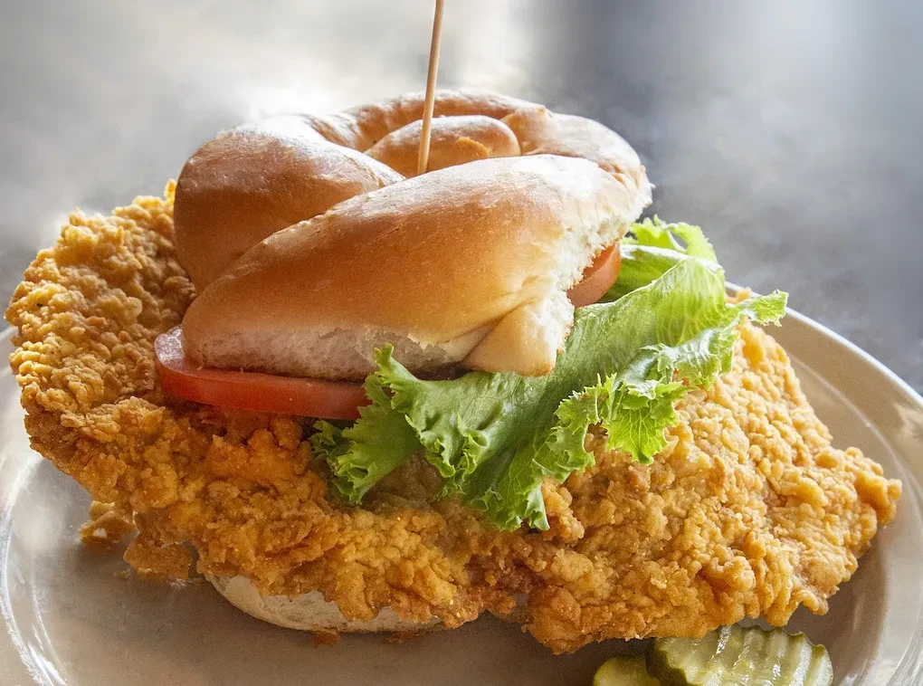 Large crispy fried chicken sandwich with lettuce, tomato, pickles, and skewer.