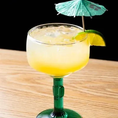 Yellow cocktail with ice, lime, and umbrella in a green-stemmed glass.