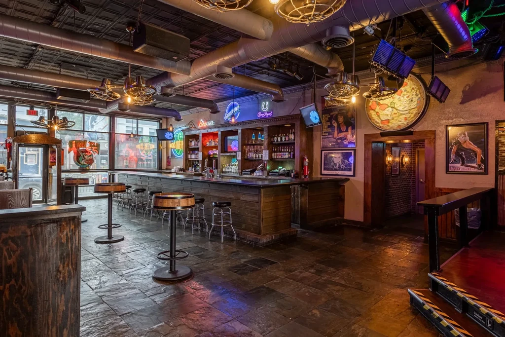 An empty industrial-eclectic bar interior with neon signs and varied art.