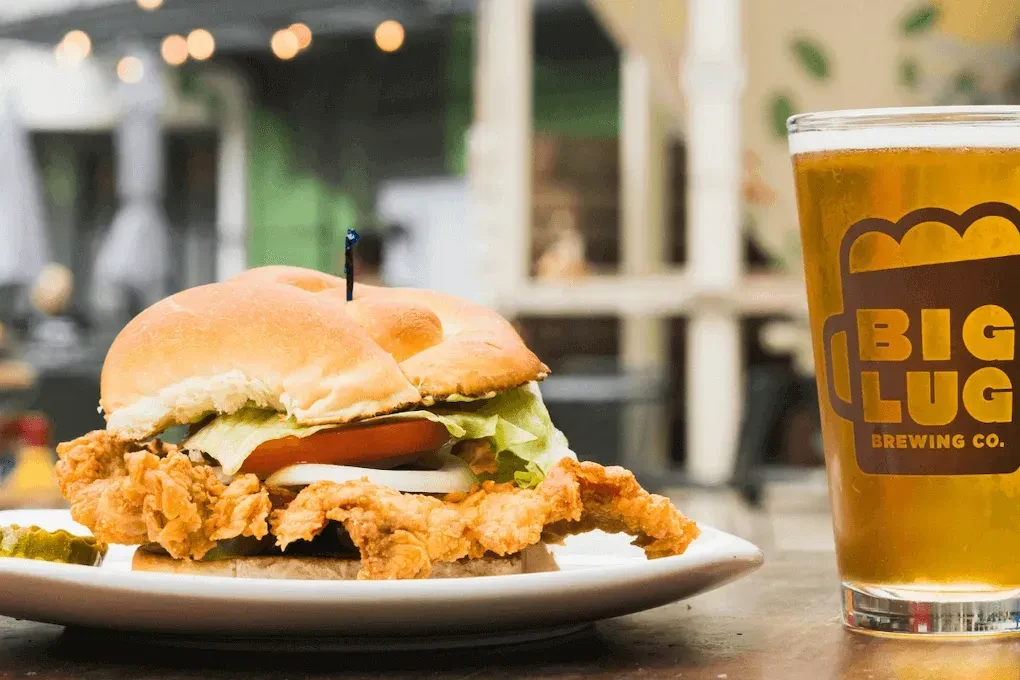 Fried chicken sandwich, pickle, and Big Lug beer glass on a wooden table.